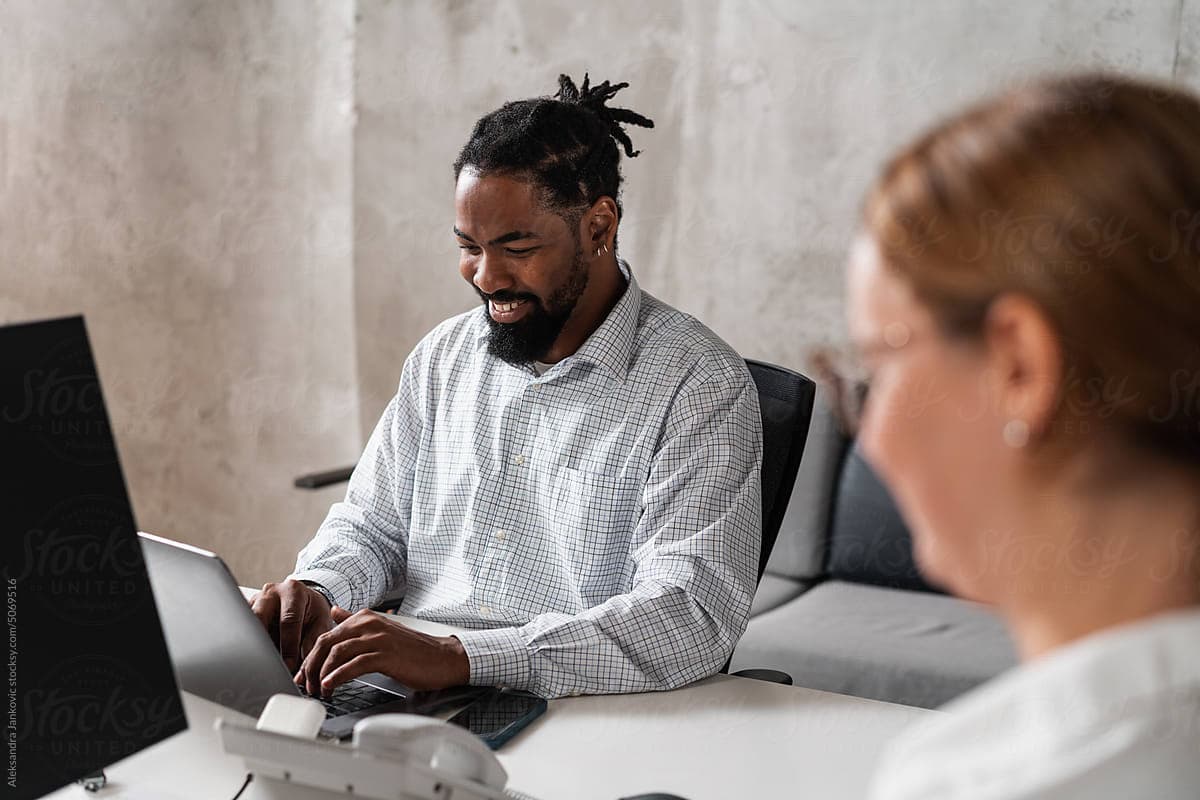 Smiling black man sitting at the desk and working on the laptop  computer. His female co-worker is anonymous and blurred.